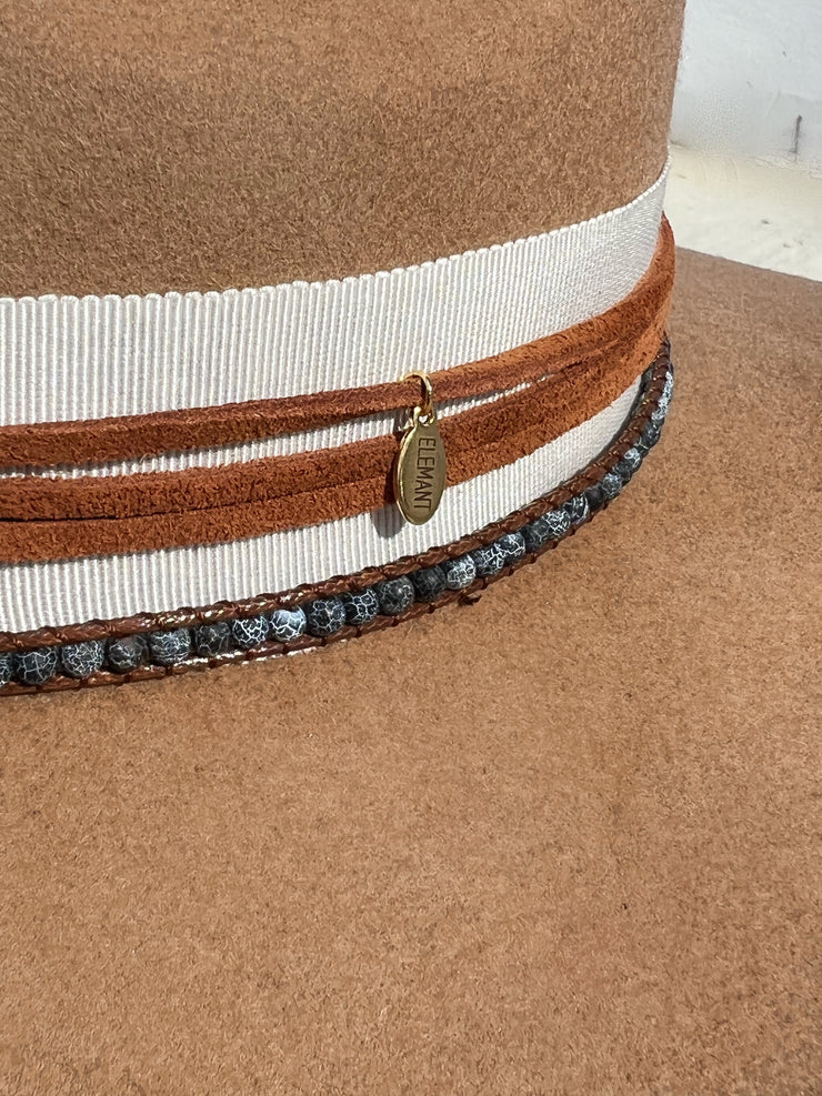 Khaki brown wool fedora hat wrapped in white hat band and strung with warm brown leather strings and a string of beads varying in colors. Natural blue druzy agate slize attached between two white and black striped feathers. Elemant gold logo emblem is hand stitched onto the top back of the hat and and another logo charm is attached and dangling on the right side of the hat from the leather string. Both visible and adding a special touch of elegance.