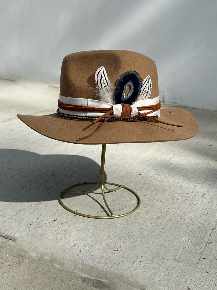 Khaki brown wool fedora hat wrapped in white hat band and strung with warm brown leather strings and a string of beads varying in colors. Natural blue druzy agate slize attached between two white and black striped feathers. Elemant gold logo emblem is hand stitched onto the top back of the hat and and another logo charm is attached and dangling on the right side of the hat from the leather string. Both visible and adding a special touch of elegance.