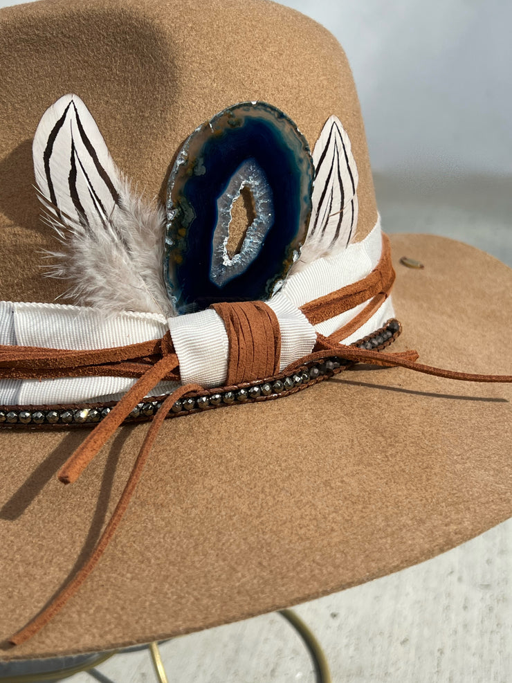 Khaki brown wool fedora hat wrapped in white hat band and strung with warm brown leather strings and a string of beads varying in colors.  Natural blue druzy agate slize attached between two white and black striped feathers.  Elemant gold logo emblem is hand stitched onto the top back of the hat and and another logo charm is attached and dangling on the right side of the hat from the leather string.  Both visible and adding a special touch of elegance.