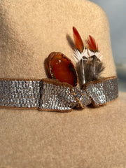 Light beige wool fedora hat with frayed metal silver band and brown underlay. Red agate stone and brown, white and orange feathers attached as well as gold Elemant metal logo on back.