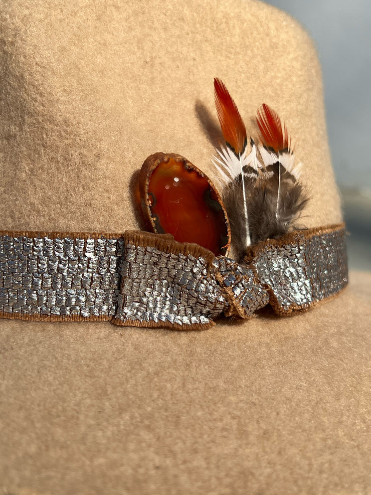 Light beige wool fedora hat with frayed metal silver band and brown underlay. Red agate stone and brown, white and orange feathers attached as well as gold Elemant metal logo on back.
