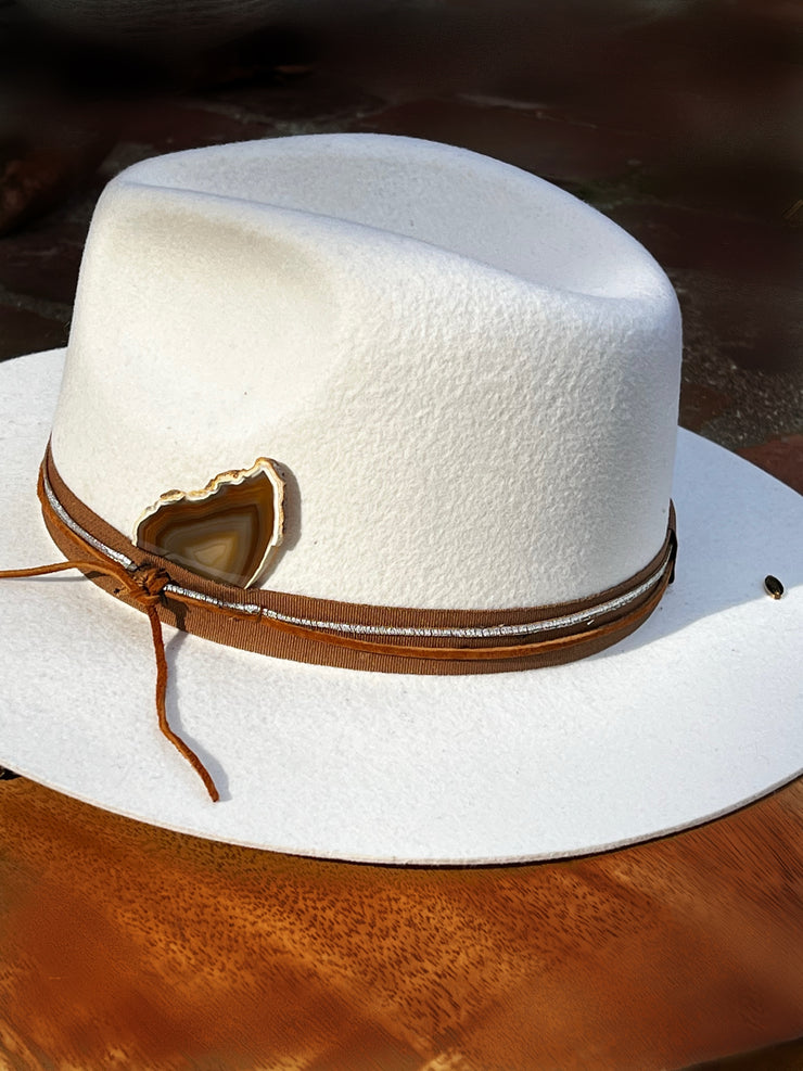 Crisp white fedora hat with brown fabric strap, leather and silver strings tied in a knot for casual yet dramatic effect. Brown agate slice with white edges and lots of color variations. Gold Elemant logo emblem attached to the top back of the hat visibly for an added touch of elegance.