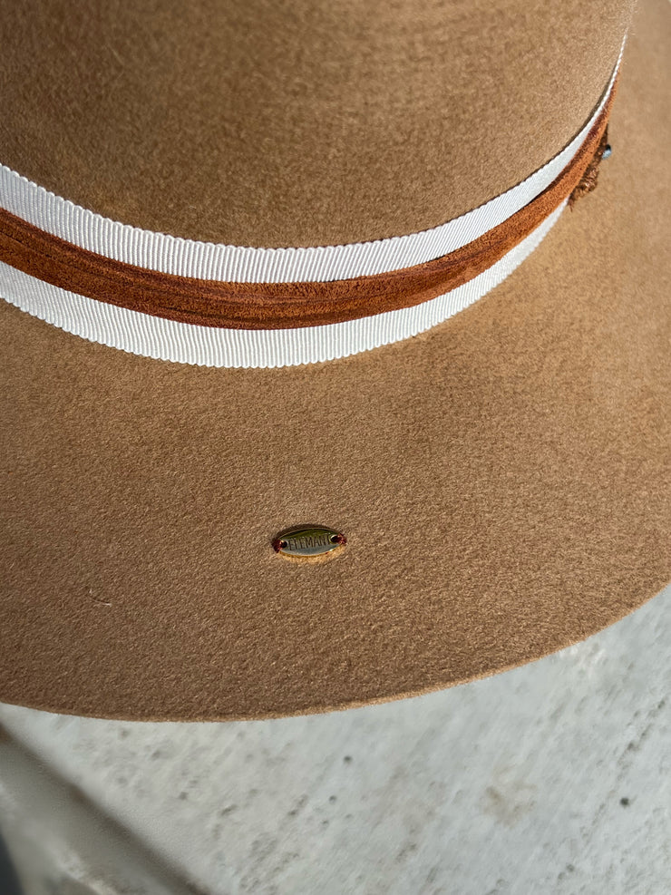 Khaki brown wool fedora hat wrapped in white hat band and strung with warm brown leather strings and a string of beads varying in colors. Natural blue druzy agate slize attached between two white and black striped feathers. Elemant gold logo emblem is hand stitched onto the top back of the hat and and another logo charm is attached and dangling on the right side of the hat from the leather string. Both visible and adding a special touch of elegance.
