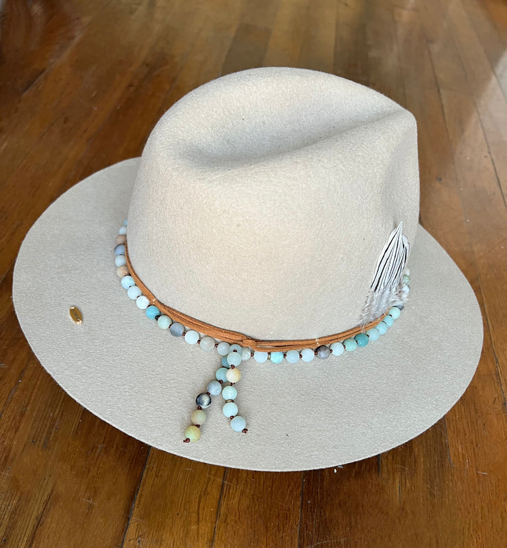 Light beige fedora hat. Light brown strings of hat belts and a string of light green, blue and brown beaded stones. Three white feathers attached to one side and a large light green agate and gold metal feather attached to the other. Gold Elemant logo stitched onto the back top of the hat brim visibly for an added touch of elegance.