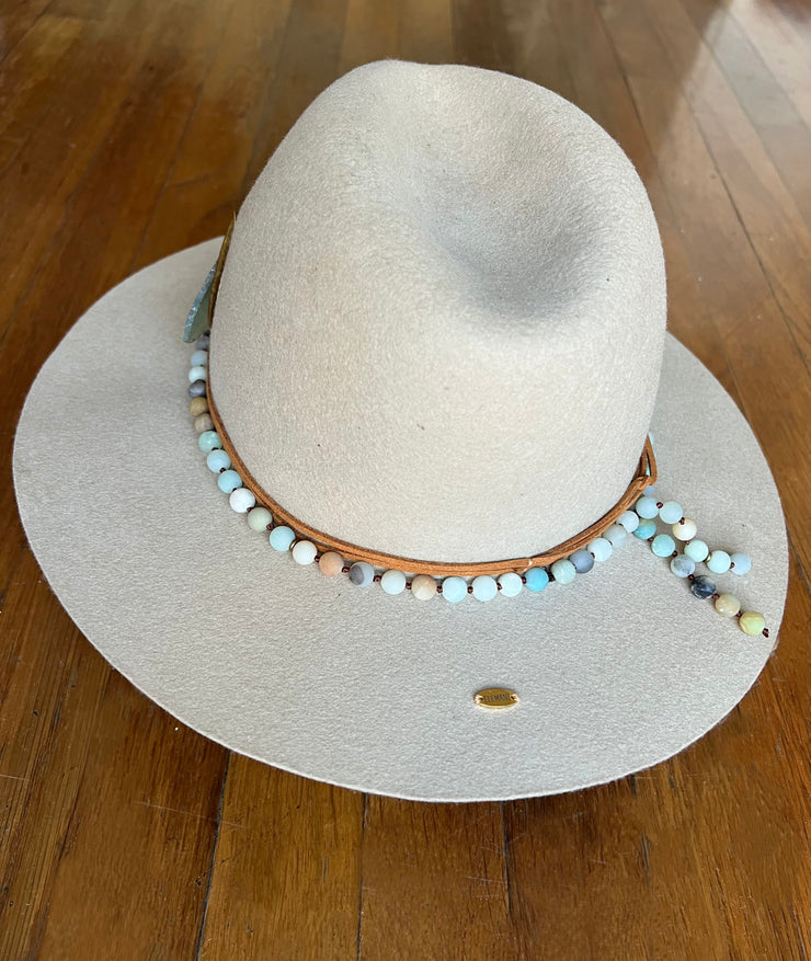 Light beige fedora hat. Light brown strings of hat belts and a string of light green, blue and brown beaded stones. Three white feathers attached to one side and a large light green agate and gold metal feather attached to the other. Gold Elemant logo stitched onto the back top of the hat brim visibly for an added touch of elegance.