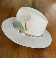 Light beige fedora hat. Light brown strings of hat belts and a string of light green, blue and brown beaded stones. Three white feathers attached to one side and a large light green agate and gold metal feather attached to the other. Gold Elemant logo stitched onto the back top of the hat brim visibly for an added touch of elegance.