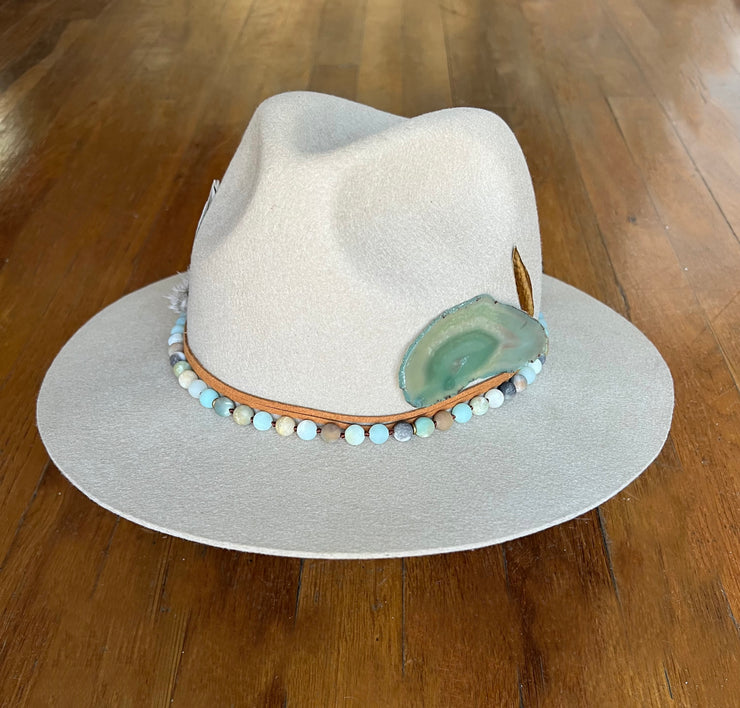 Light beige fedora hat.  Light brown strings of hat belts and a string of light green, blue and brown beaded stones.  Three white feathers attached to one side and a large light green agate and gold metal feather attached to the other.  Gold Elemant logo stitched onto the back top of the hat brim visibly for an added touch of elegance.