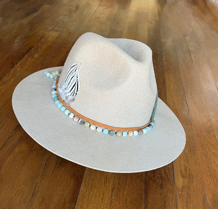 Light beige fedora hat. Light brown strings of hat belts and a string of light green, blue and brown beaded stones. Three white feathers attached to one side and a large light green agate and gold metal feather attached to the other. Gold Elemant logo stitched onto the back top of the hat brim visibly for an added touch of elegance.