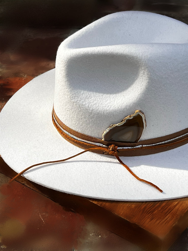 Crisp white fedora hat with brown fabric strap, leather and silver strings tied in a knot for casual yet dramatic effect.  Brown agate slice with white edges and lots of color variations.  Gold Elemant logo emblem attached to the top back of the hat visibly for an added touch of elegance.
