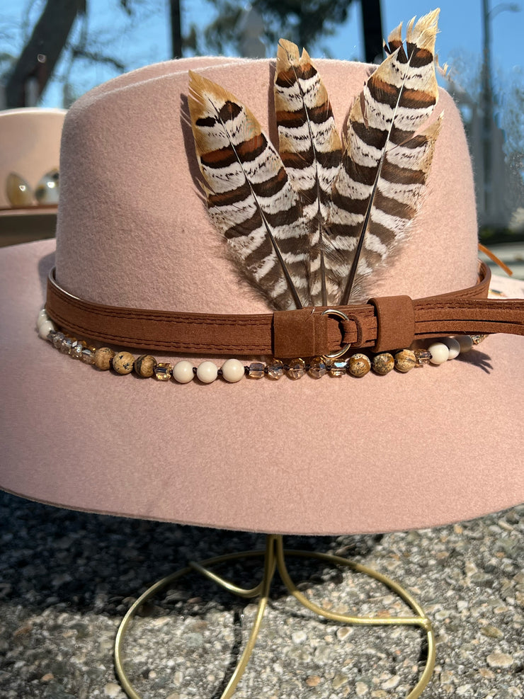 Dusty pink wool fedora hat.  Brown belt and buckle hat band as well as a string of beads and stones.  Three large feathers attached on one side and two natural white, brown speckled, agate stones on the other.  Gold Elemant logo emblem stitched on to the back top of the hat brim visibly for an added touch of elegance.
