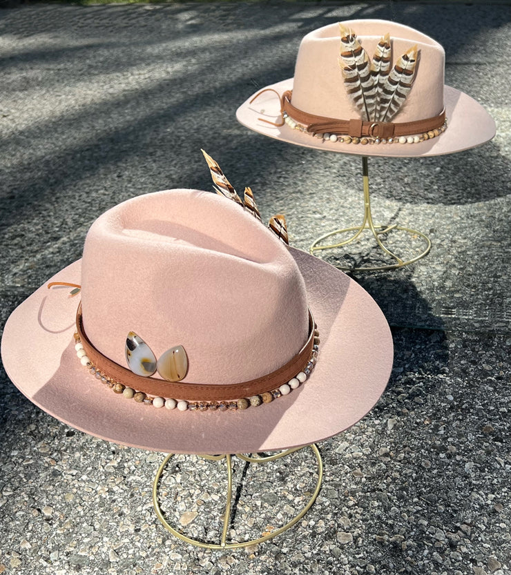 Dusty pink wool fedora hat. Brown belt and buckle hat band as well as a string of beads and stones. Three large feathers attached on one side and two natural white, brown speckled, agate stones on the other. Gold Elemant logo emblem stitched on to the back top of the hat brim visibly for an added touch of elegance.