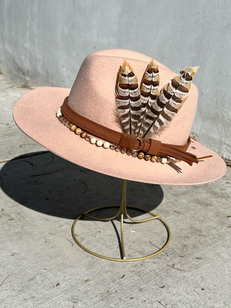 Dusty pink wool fedora hat. Brown belt and buckle hat band as well as a string of beads and stones. Three large feathers attached on one side and two natural white, brown speckled, agate stones on the other. Gold Elemant logo emblem stitched on to the back top of the hat brim visibly for an added touch of elegance.