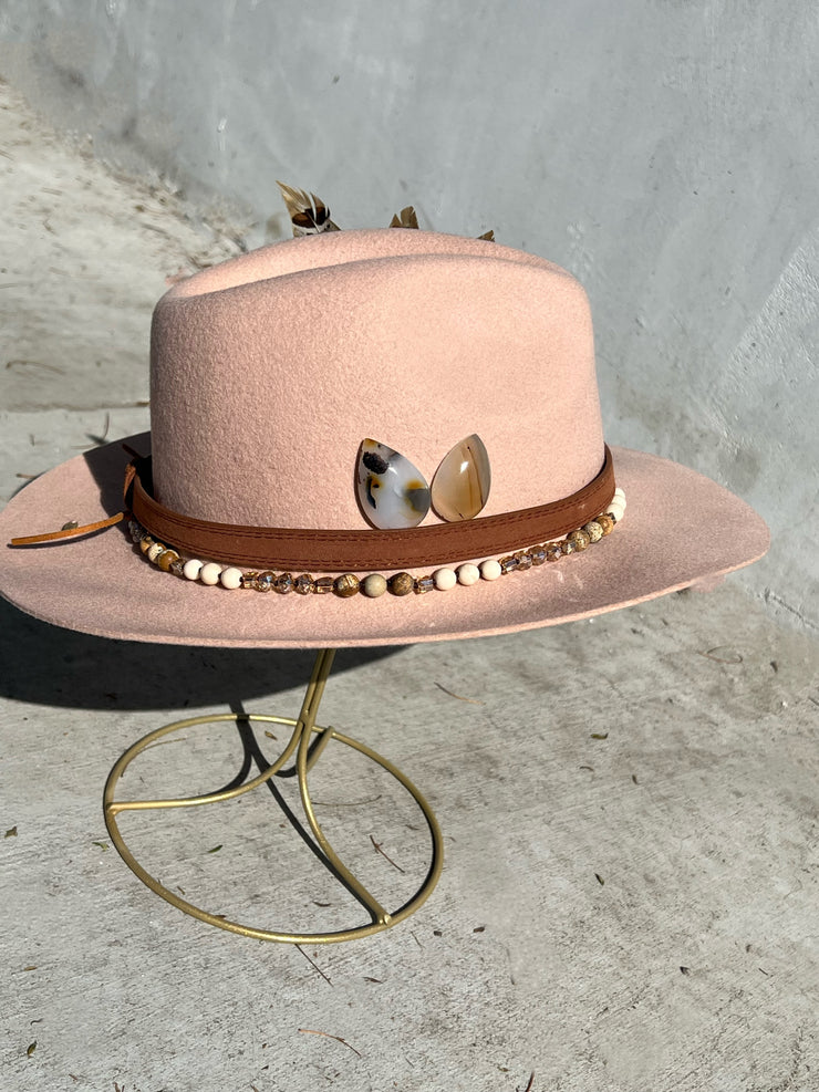 Dusty pink wool fedora hat. Brown belt and buckle hat band as well as a string of beads and stones. Three large feathers attached on one side and two natural white, brown speckled, agate stones on the other. Gold Elemant logo emblem stitched on to the back top of the hat brim visibly for an added touch of elegance.